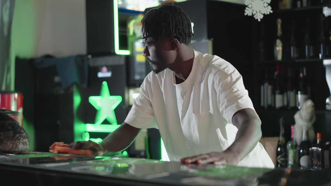 Bartender preparing drinks at bar counter with focused expression. Snowflake decorations hang above. Behind him, stocked shelves with bottles, including festive Heineken displays