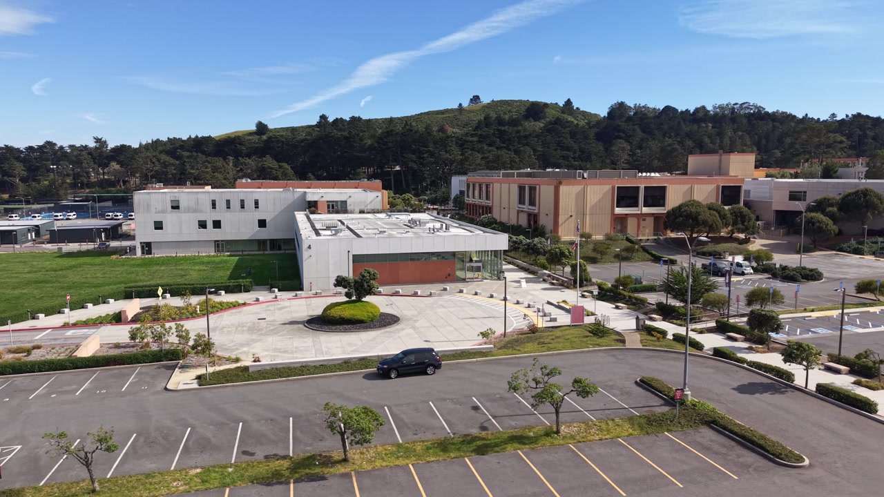 Aerial view of the modern architectural buildings and the nearly empty parking lot of Skyline College in San Bruno, California, on a sunny day with hills in the background