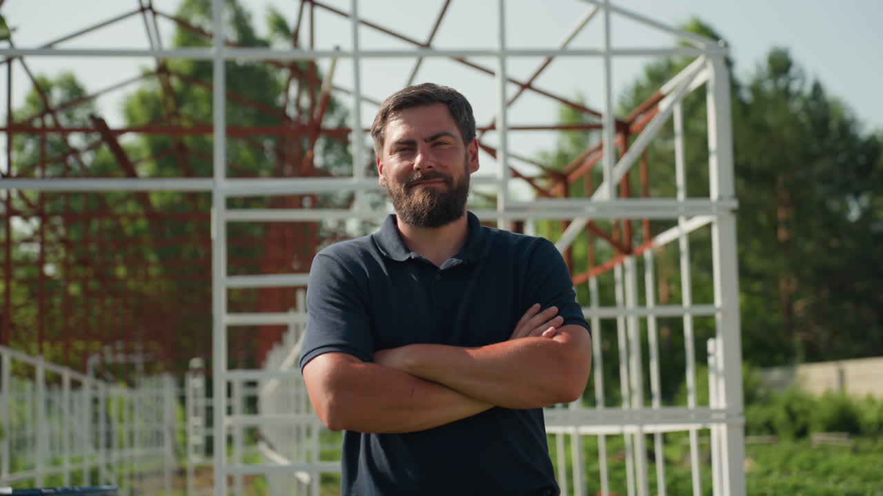 Trabajador de rancho girando con las manos cruzadas junto a un edificio de estructura metálica, con expresión tranquila, inspeccionando el progreso de la propiedad rural, con la luz del sol sobre la vegetación y la estructura en un sitio agrícola al aire libre.