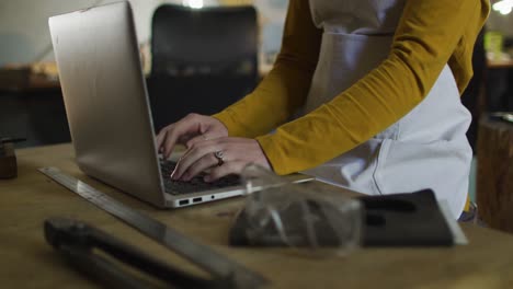 Midsection-of-caucasian-female-jeweller-in-workshop-wearing-apron,-sitting-at-desk,-using-laptop