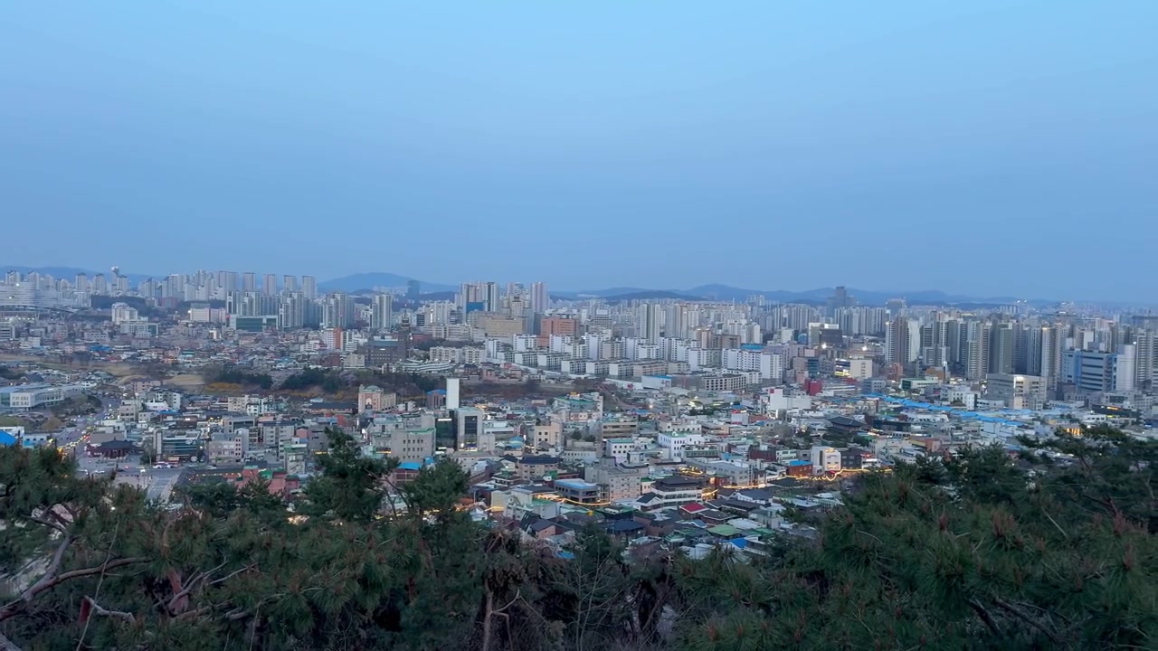 Pemandangan panorama kota Suwon saat senja, menampilkan lingkungan perumahan, gedung-gedung tinggi modern, dan latar belakang pegunungan yang jauh.
