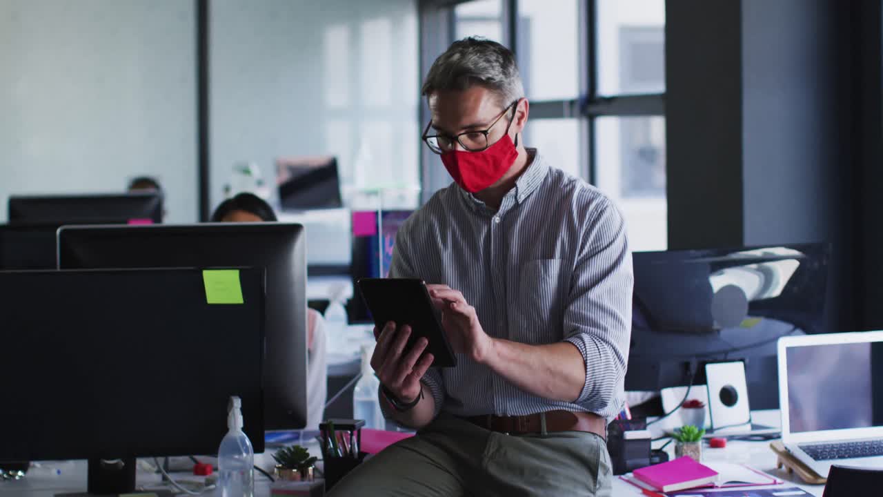 Caucasian man wearing face mask using digital tablet while sitting over his desk at modern office