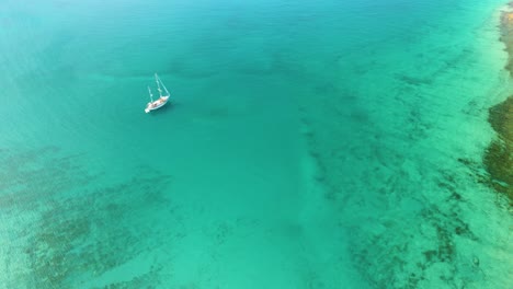 Imágenes-Aéreas-De-Un-Velero-Blanco-En-El-Tranquilo-Mar-Mediterráneo-Frente-A-Una-Isla-Paradisíaca-Con-Mar-Azul-Turquesa-Y-Exuberante-Vegetación-Verde-Vista-Aérea