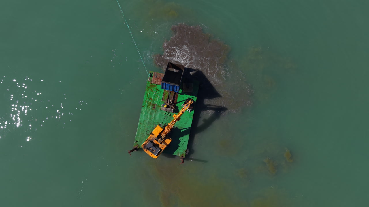 Top-down view of a green dredging barge with yellow excavator working in shallow turquoise water, stirring up sediment plumes near a tow cable