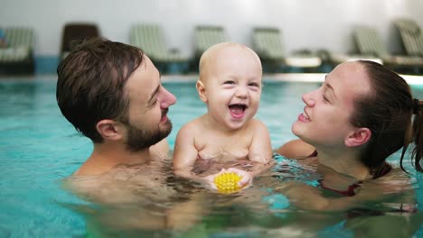 Young-mother-and-father-are-holding-their-cute-child-in-their-hands-in-the-swimming-pool-and-kissing-him-from-both-sides.-Happy-child-is-smiling