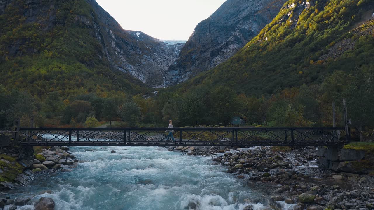 niña caminando sobre un puente que cuelga sobre un río salvaje con rocas a los lados, paisaje de montaña en el fondo