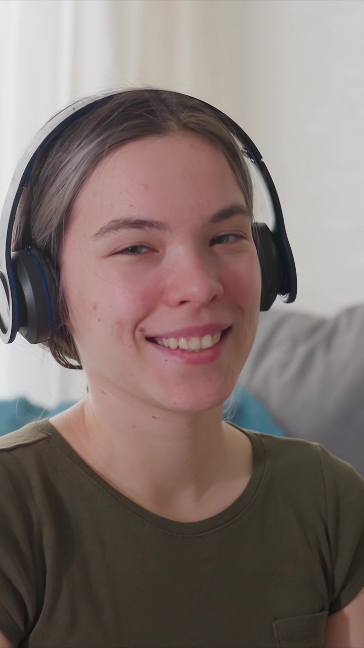 Una giovane donna indossa cuffie nere over-ear in una morbida luce naturale. La donna rimane immobile, sorridendo leggermente con gli occhi aperti o chiusi a intermittenza su uno sfondo interno sfocato