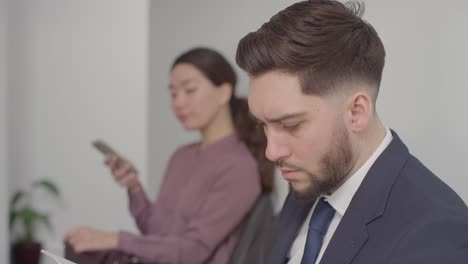 male and female candidates in office waiting for job interview looking at mobile phone and reading notes 2