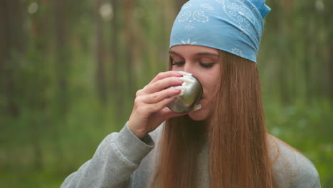 close-up of young woman with blue bandana sipping water from thermos cup in serene green forest, focusing on her calm and relaxed expression, with delicate, well-manicured nails