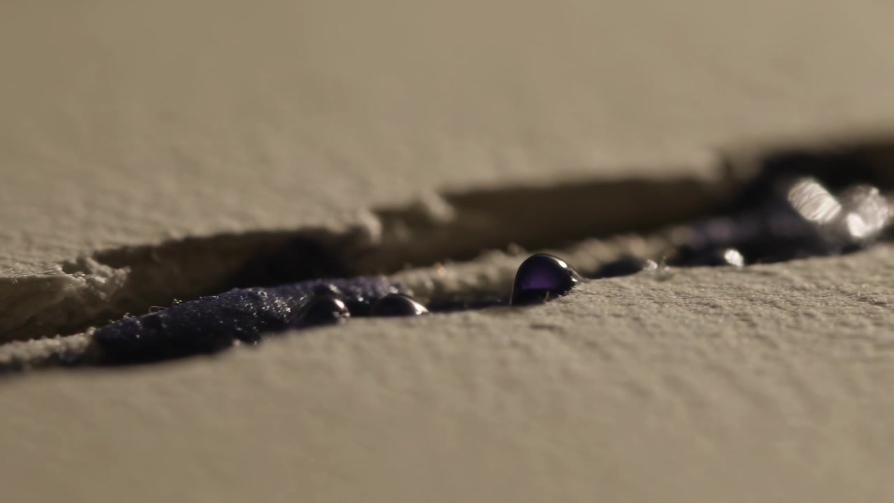 Thin purple film retracting into glossy droplets along beige paper groove, lit by warm angled light