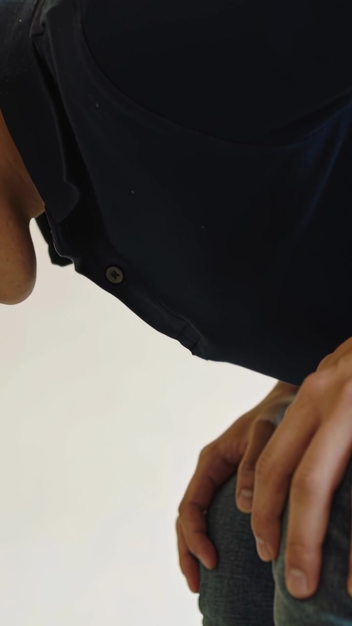 Vertical video: Leaning man reacting to low angle indoors, dark shirt, jeans, hands on right knee