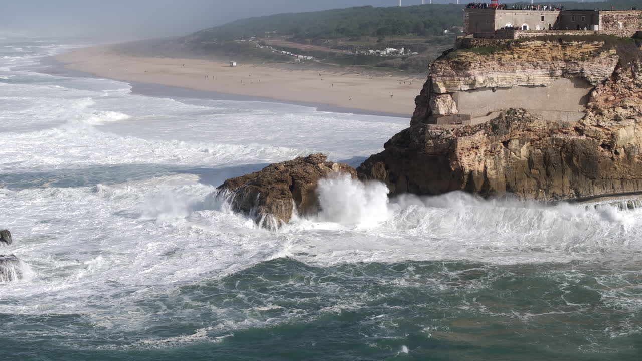 Aerial drone shot of big waves breaking on rocks and cliffs on a day with giant waves in Nazaré, Portugal, Europe. Farol da Nazaré lighthouse and wind turbines visible. Drone slowly orbiting
