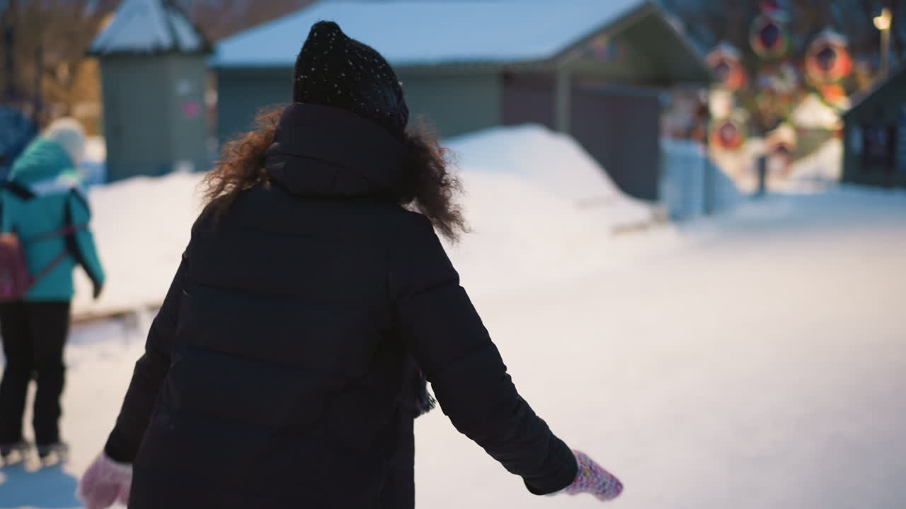 Skater in black jacket, knitted hat, and colorful mittens glides outdoors on snowy winter path during evening with festive lights in background, arms spread for balance, enjoying seasonal cold atmosphere