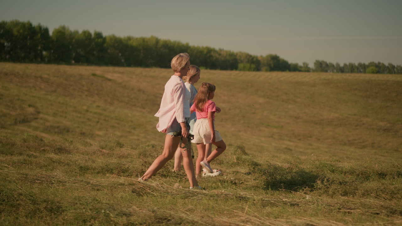 Side view of child walking with her two aunts through vast open field, one aunt holding rope while they enjoy outdoor stroll, surrounded by nature, lush green trees in background