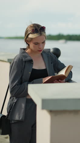 mujer con ropa gris descansa su mano derecha en una valla de hierro mientras lee un libro a la orilla del río, el viento sopla suavemente las páginas del libro, con árboles lejanos y agua visibles en el fondo