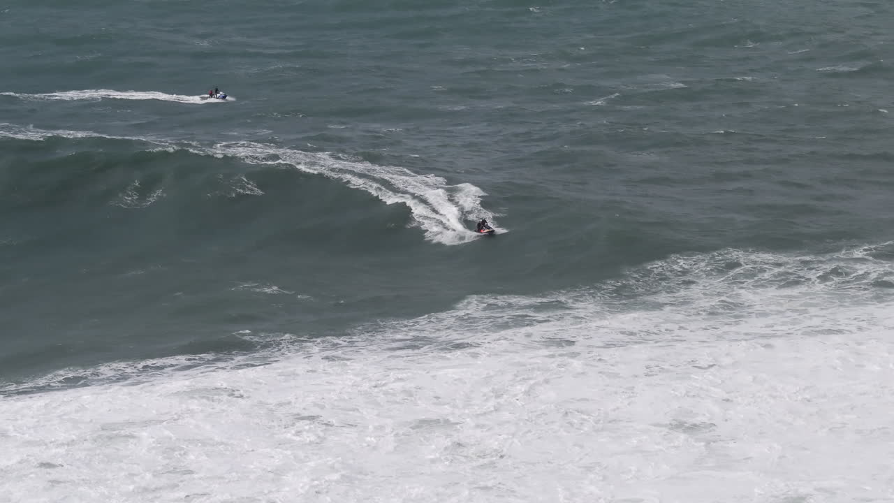 Jet ski riders moving through enormous Nazaré surf aerial view. Portugal, Europe, big wave tow-in professional surfing