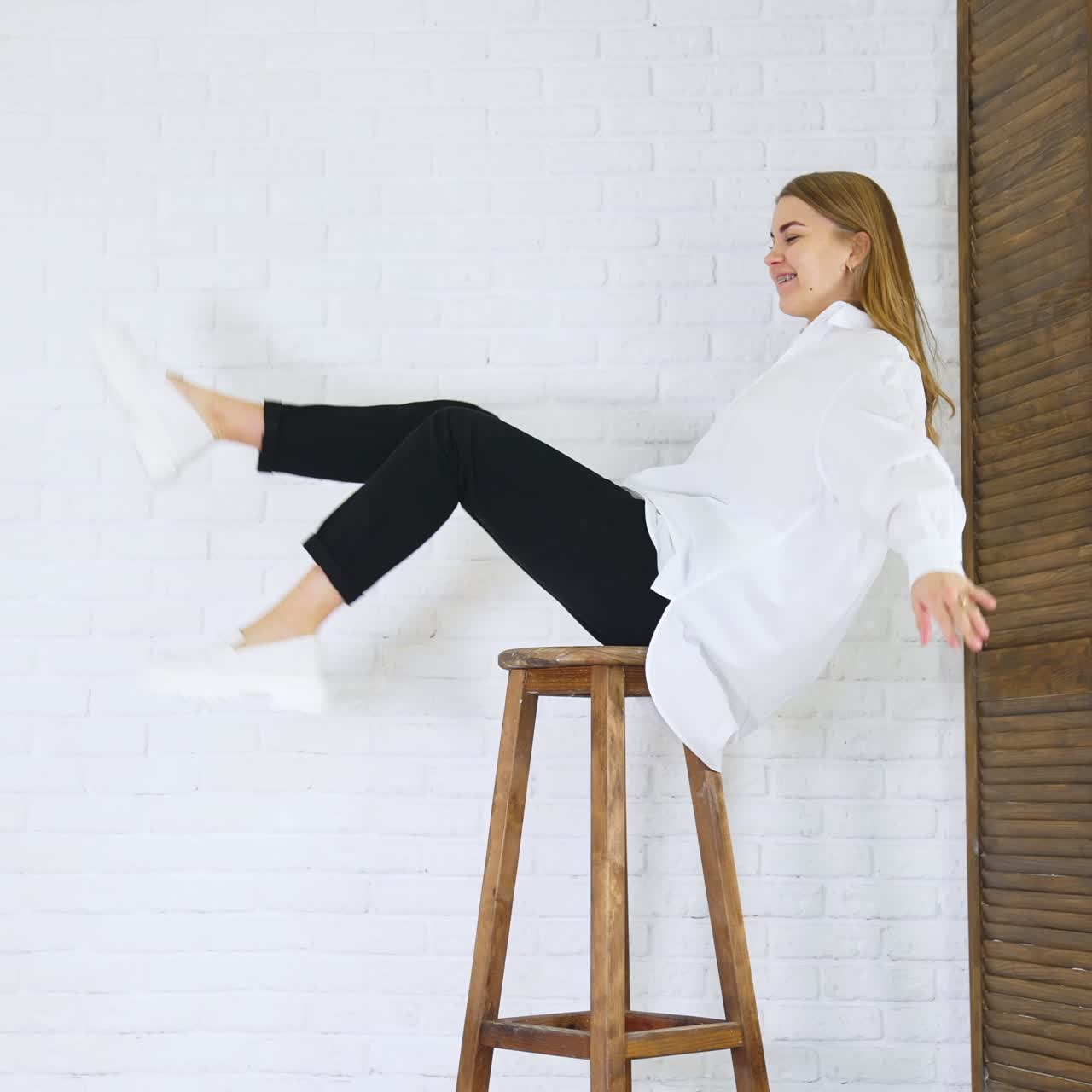 Long-haired lady in white shirt sits on a high stool waving her legs. White modern shoes presentation at the backdrop of white brick wall