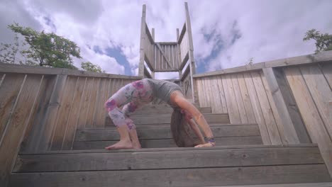 Beautiful-young-woman,-performing-wheel-pose-on-wooden-stairs,-outdoors