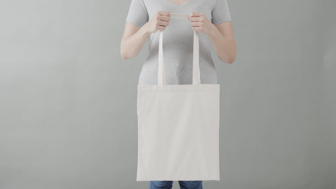 mujer caucásica con una camiseta blanca sosteniendo una bolsa blanca sobre un fondo gris, espacio de copia, cámara lenta