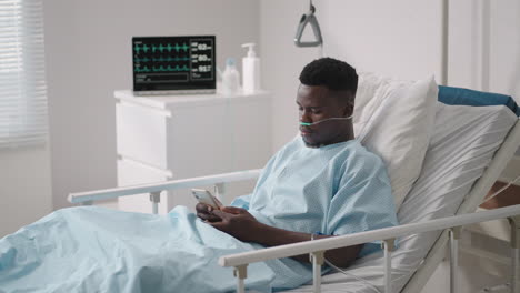 Paciente-Varón-Africano-Usando-Un-Teléfono-Inteligente-Mientras-Está-Acostado-En-La-Cama.-Paciente-Africano-Teniendo-Una-Conversación-Telefónica-En-La-Habitación-Del-Hospital.
