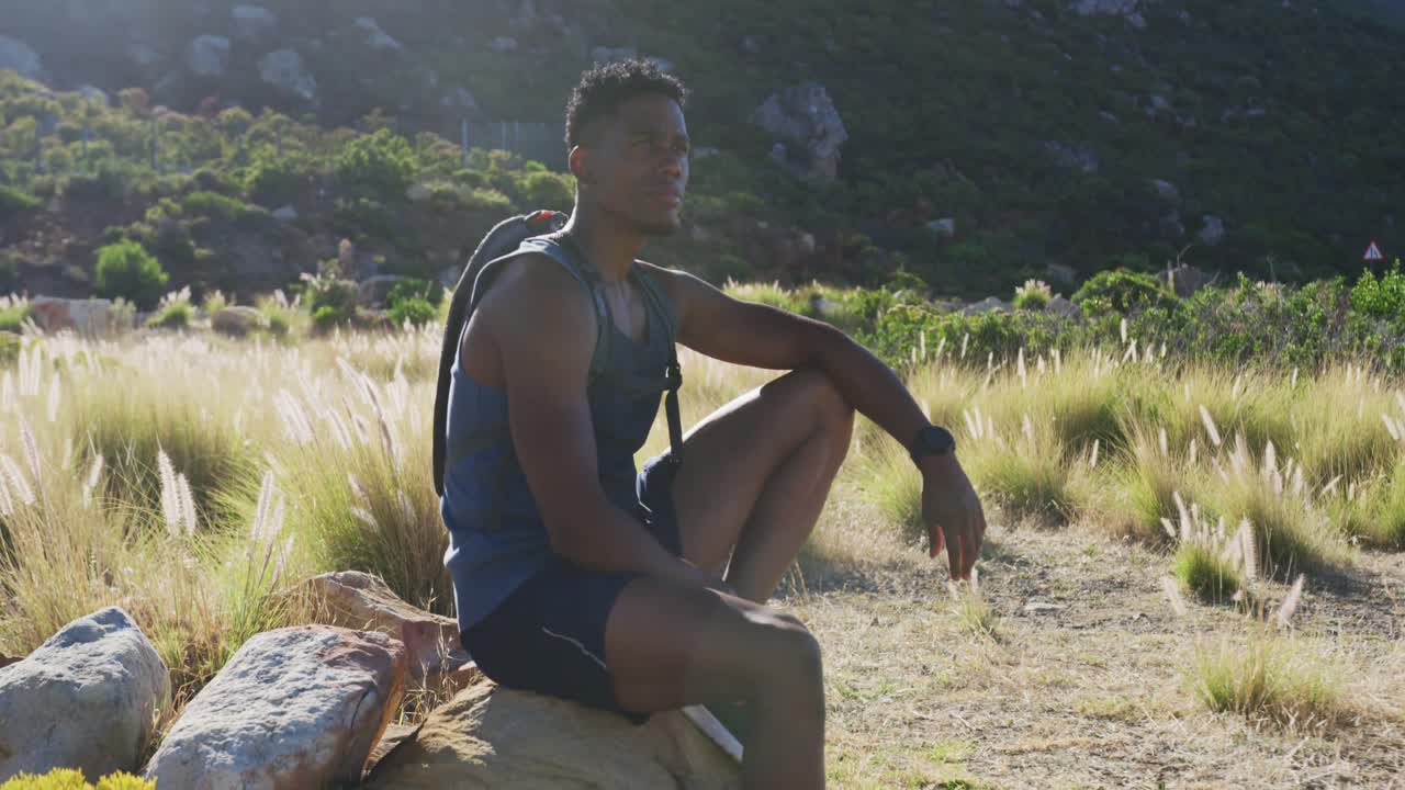 Uomo afroamericano che si allena all'aperto, fa trekking e riposa su una roccia in un paesaggio montano.
