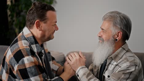 From-the-side,-a-happy-brunette-with-stubble-in-a-plaid-shirt-holds-the-hands-of-his-elderly-boyfriend-with-gray-hair-and-a-full-beard,-and-they-press-their-foreheads-to-each-other,-looking-into-the-eyes-while-sitting-on-the-sofa-in-a-modern-apartment