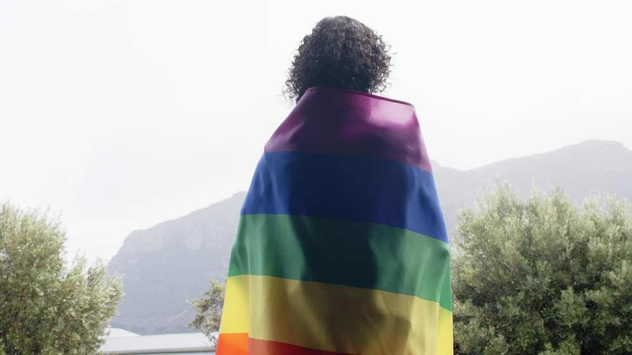 Pessoa não binária avistando a vista da montanha, ajustando a bandeira do arco-íris, desfraldando-a para mostrar orgulho no terraço.