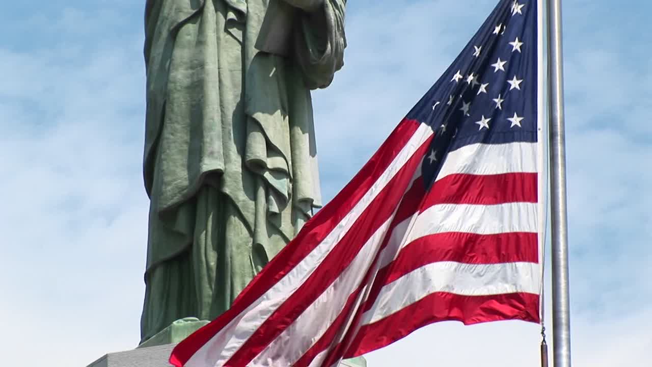 la estatua de la libertad se erige majestuosamente contra un cielo azul lleno de nubes tenues mientras una bandera estadounidense ondea en primer plano