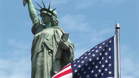 Majestätisch-Steht-Die-Freiheitsstatue-Vor-Einem-Blauen-Himmel-Voller-Wolkenfetzen,-Während-Im-Vordergrund-Eine-Amerikanische-Flagge-Weht