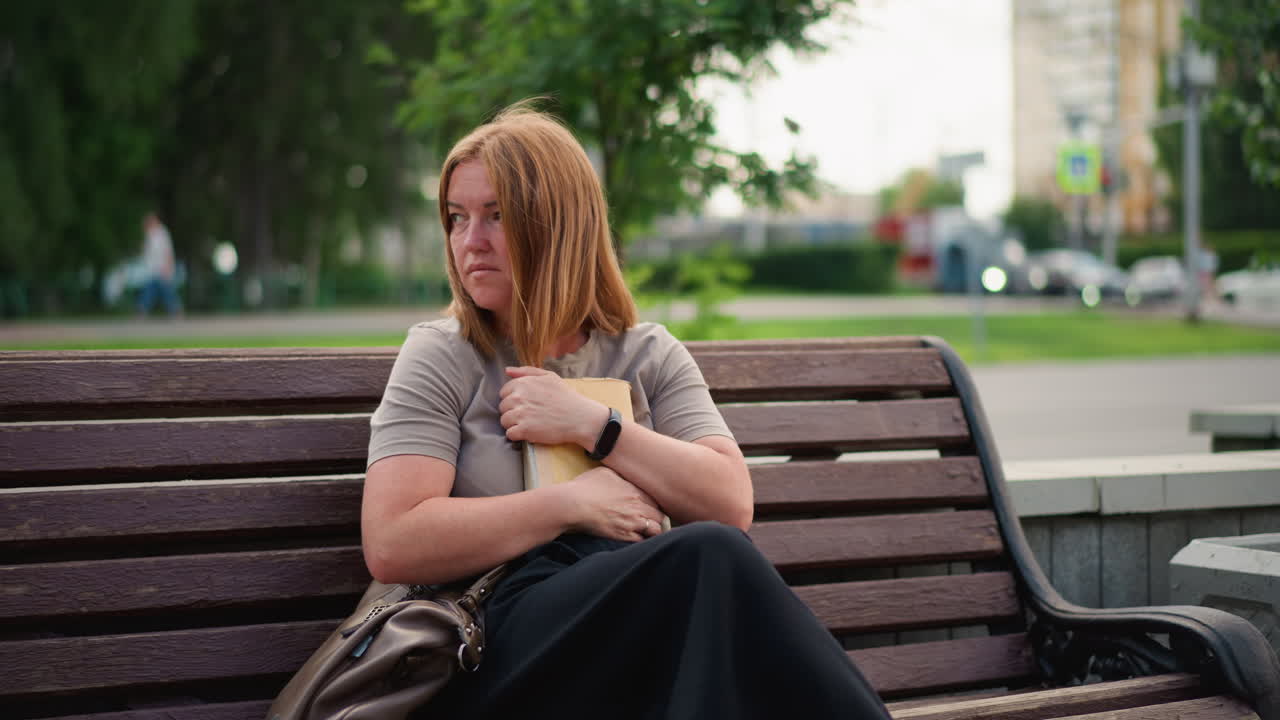 Unhappy woman sits alone on wooden bench outdoors closing book and pressing it to chest, thoughtful expression suggesting sadness or reflection, cars and greenery blurred