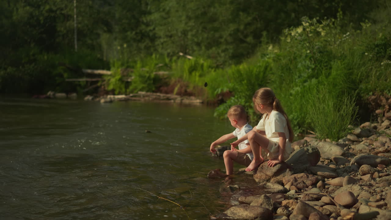 Girl and boy throw stones into river playing together in nature. Spending summer holidays with family in tourist place without using gadgets for children slow motion
