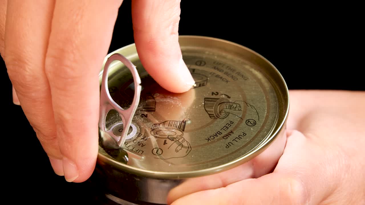 A hand opens a metal can using a pull tab, shown in a close-up sequence with bright lighting and a black background, emphasizing the action