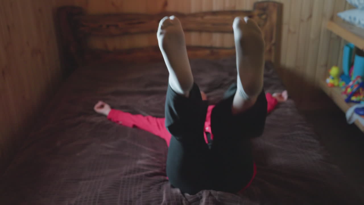 lady in red top smiles joyfully while sitting on bed in wooden room then gently falls backward on bed playfully showing carefree moment filled with happiness in cozy indoor rustic setting