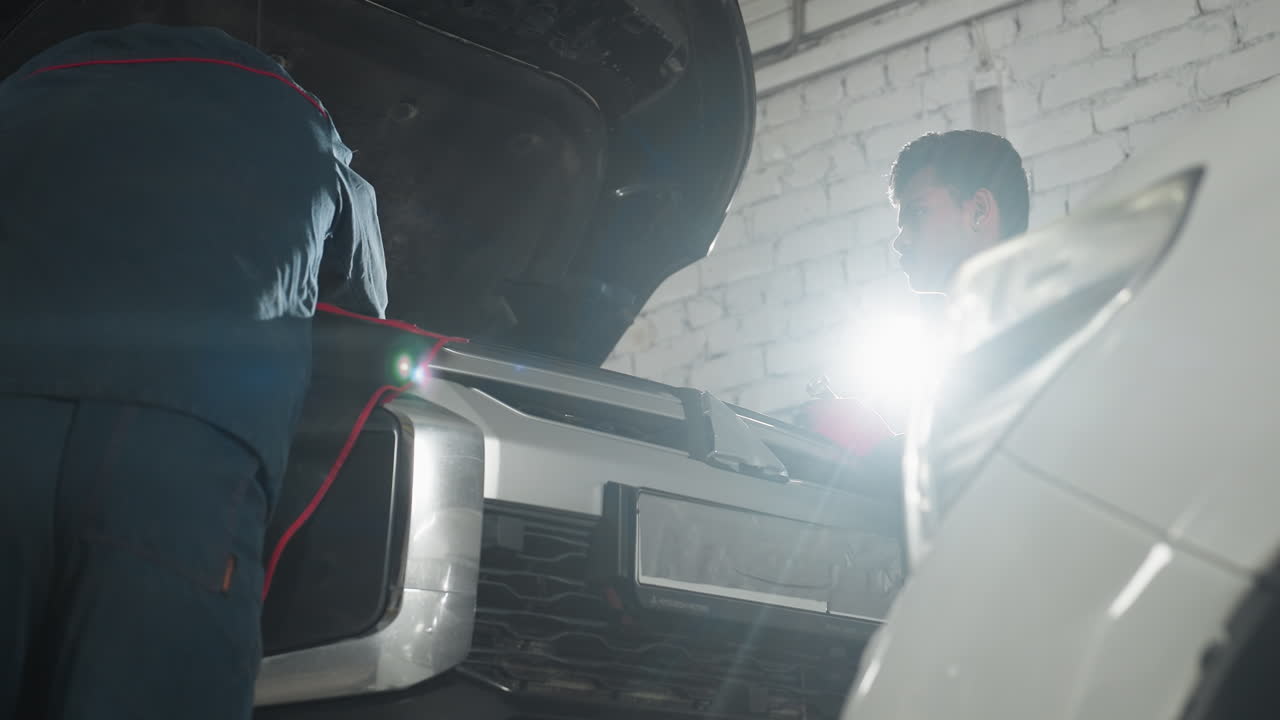 두 명의 메카닉이 오토 워크에서 검은 차를 고치고 있습니다. 하나는 엔진을 고치고 다른 하나는 관찰합니다. 강렬한 백라이트는 실루 효과를 만니다.