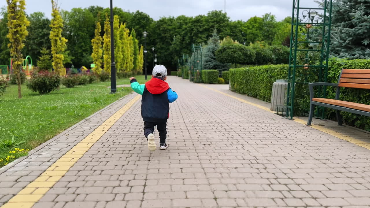Happy kid in a jacket, cap and pants running by the park. Following the cheerful kid having active time outdoors.