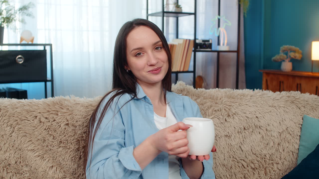 Young woman comfortably sitting on sofa drinking aromatic tea or coffee enjoying cozy calm morning