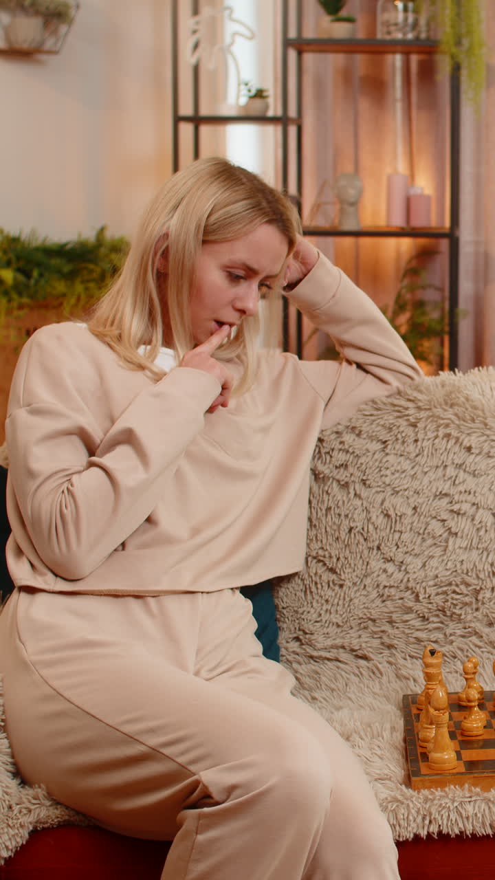 Adult woman playing chess alone at home on sofa focusing making thoughtful move with deep expression