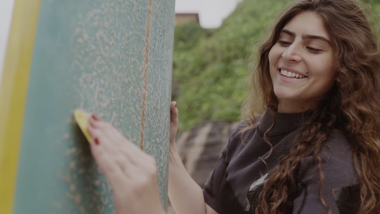 A smiling woman waxing her surfboard before a surf session