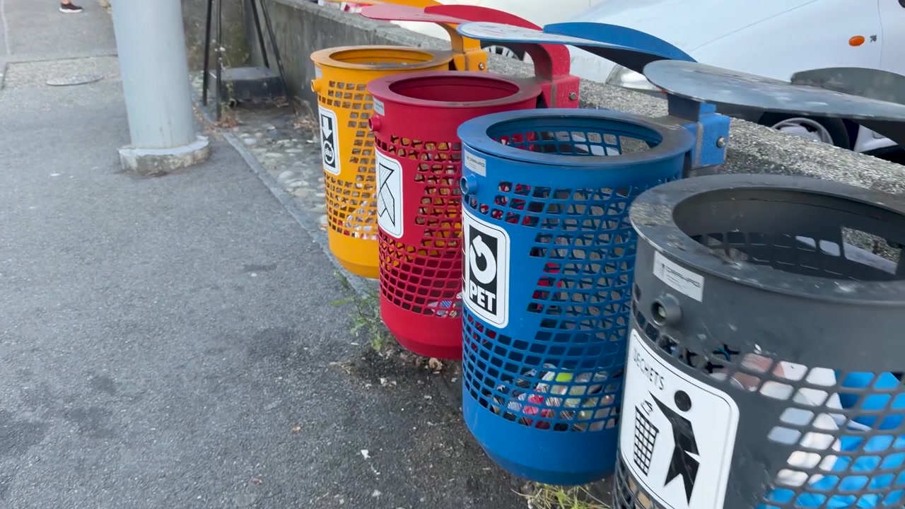 Camera pans across a row of color-coded metal recycling bins on a city sidewalk.