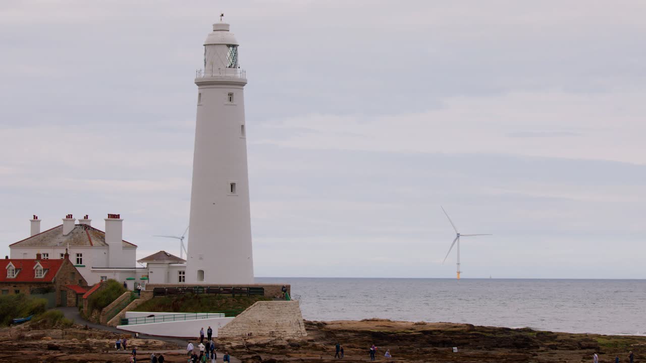 Wide static shot of a white lighthouse, adjacent cottages, and distant wind turbines along the Whitley Bay coastline under soft, diffused daylight