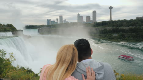 pareja multiétnica de pie en un abrazo en la plataforma de observación de las cataratas del niágara mirando hacia el di