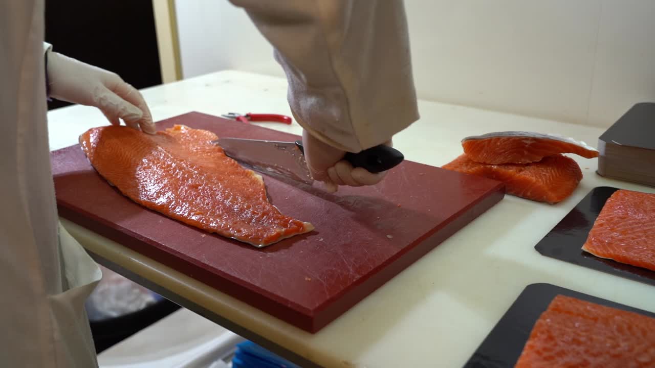 Fish industry worker in white coverall is cutting salmon fillet on plastic chopboard - Static handheld slow motion from behind