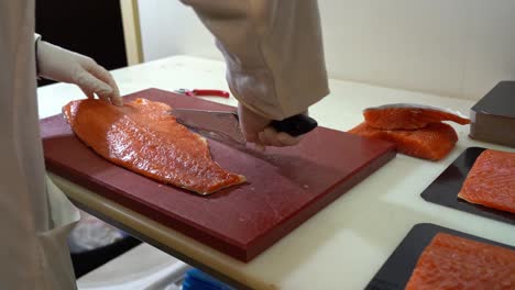 fish industry worker in white coverall is cutting salmon fillet on plastic chopboard - static handheld slow motion from behind