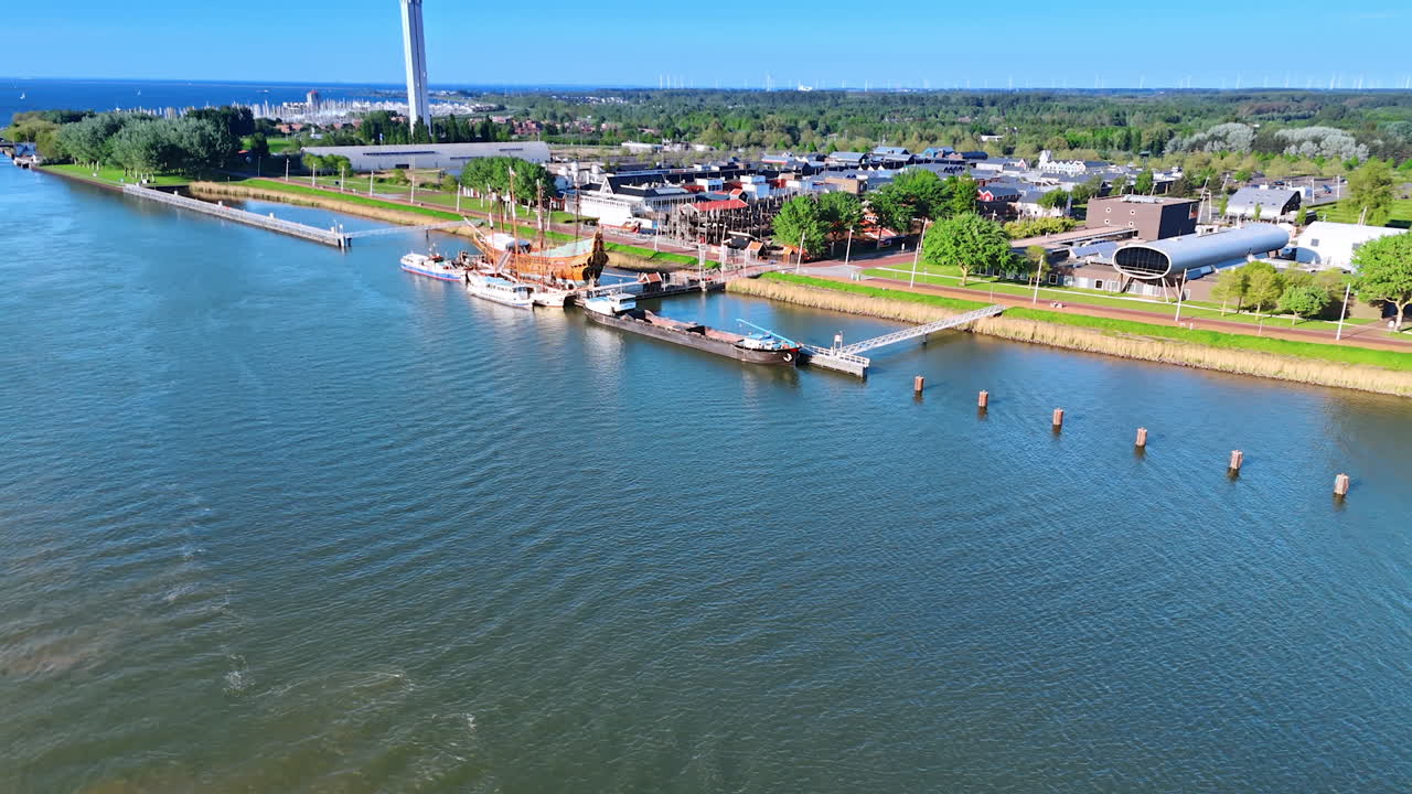 Moving over the lake waterscape approaching the waterfront. Reconstruction of the ship Batavia near the berth. Telecom tower at backdrop. Aerial perspective.