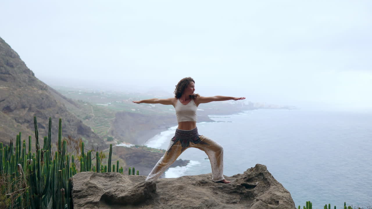 The woman's yoga warrior pose at the ocean, beach, and rocky mountains not only reflects motivation and inspiration but also emphasizes a wholesome fitness lifestyle amidst nature's beauty