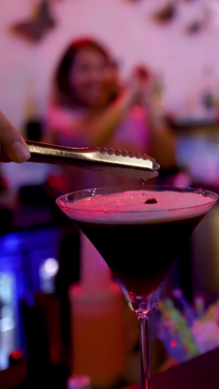 Un barista aggiunge una guarnizione a un espresso martini sotto l'illuminazione colorata del bar, con una profondità di campo ridotta.