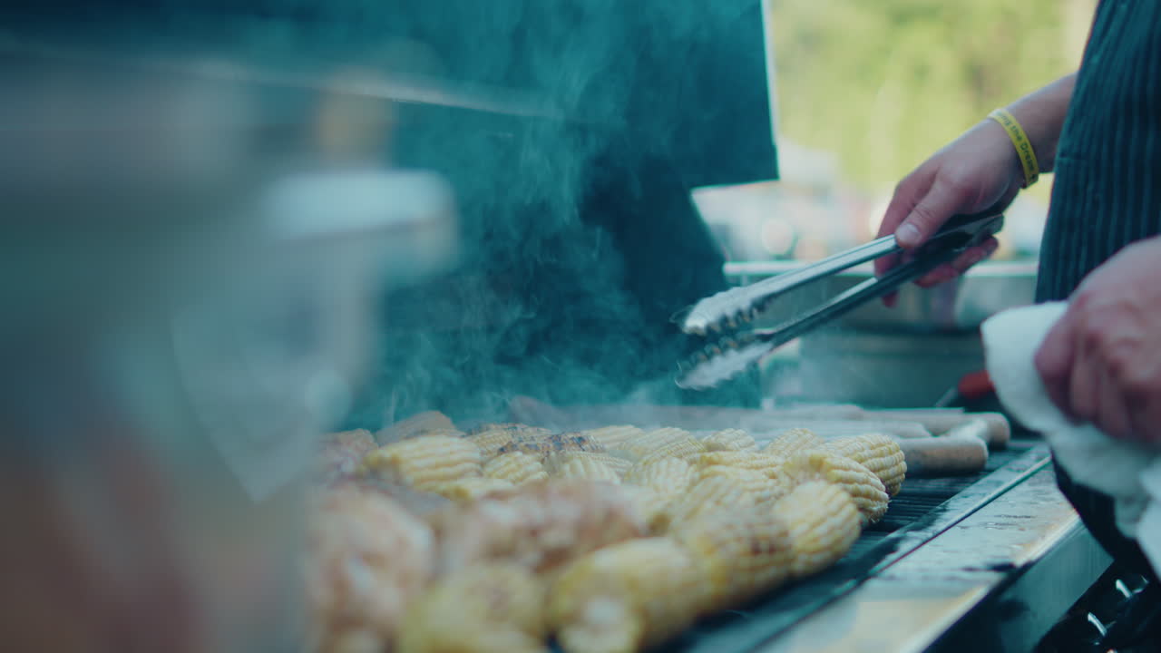 Smoking BBQ grill filled with corn, as a man skillfully uses metal tongs to turn them around. The vibrant smoke and create an inviting outdoor cooking atmosphere.