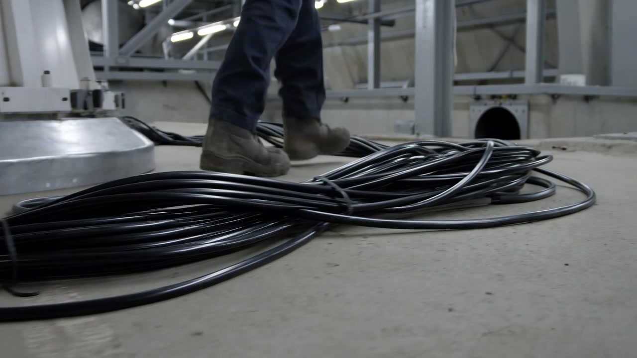 Industriel lavvinkeludsigt af en arbejder, der går forbi oprullede elektriske kabler på et betongulv ved siden af stålkonstruktion, forsyningsudstyr og overliggende støtter inde i et dæmpet servicelokale.