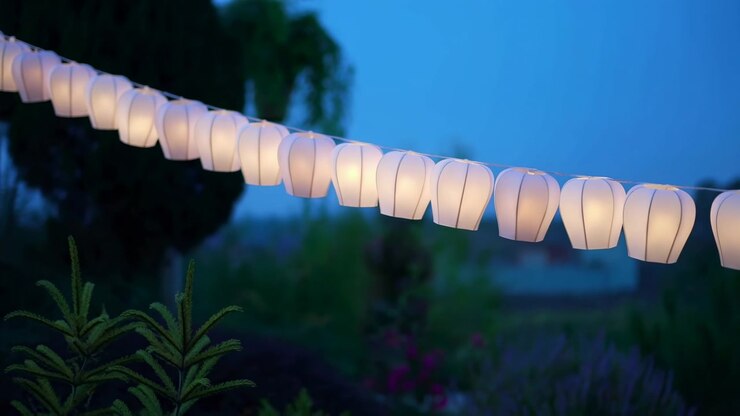 Row of white paper lanterns lit with soft creamy glow suspended on taut string across outdoor garden at dusk