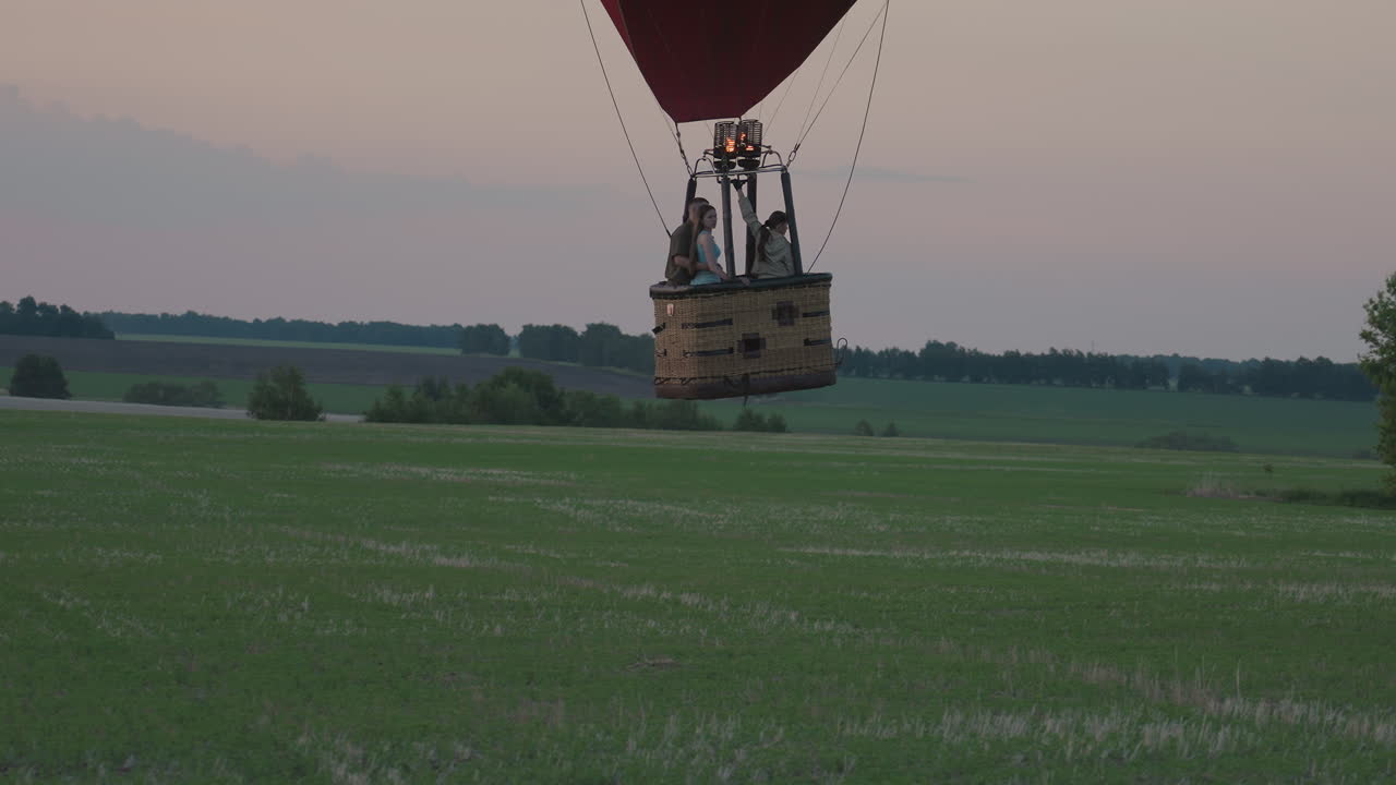 three passengers ride in hot air balloon basket over green farmland at dusk with flame igniting from burner creating serene aerial adventure under pastel sunset sky